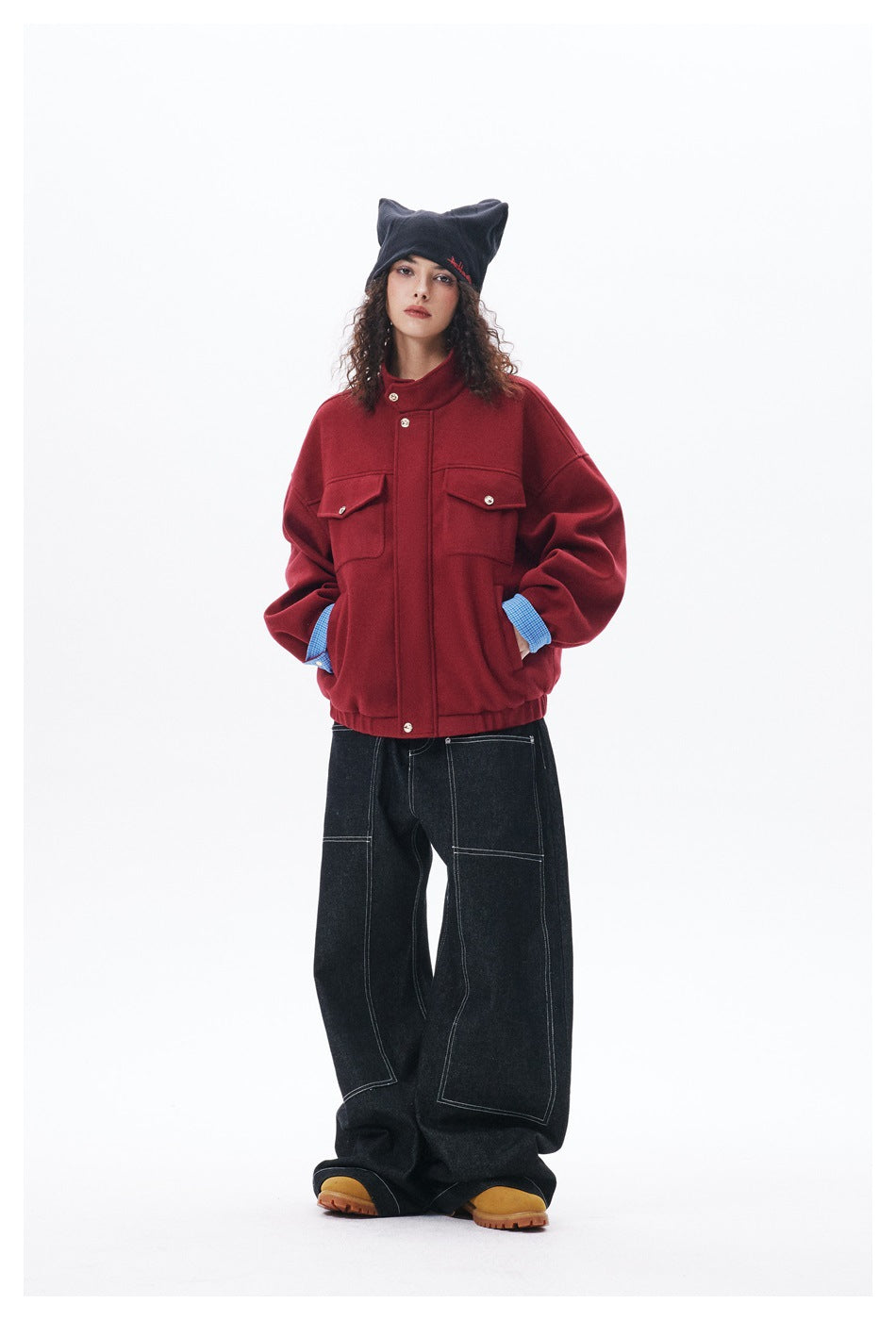 Model poses front-facing in a red jacket with checked cuffs and black wide-leg jeans, wearing a black pointed beanie.