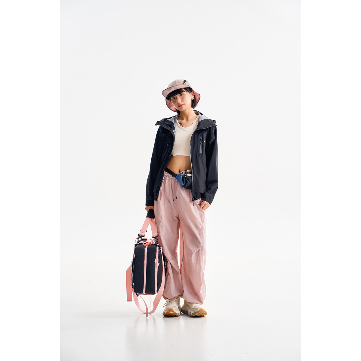 Model wearing a dark hooded jacket, white crop top, and loose pale pink pants against a white backdrop.