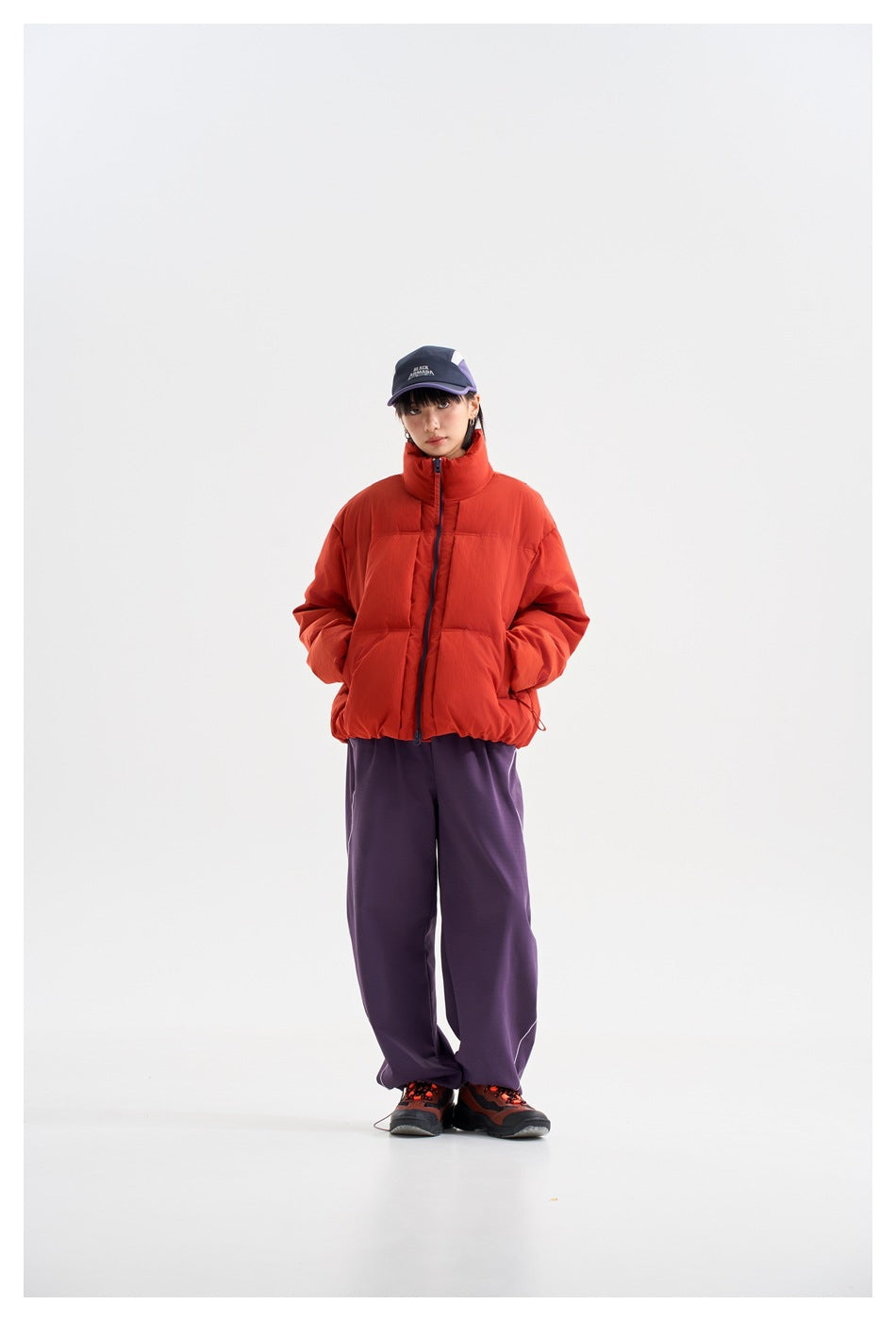 Front view of a model wearing a red-orange puffer jacket with navy zipper and loose purple trousers.
