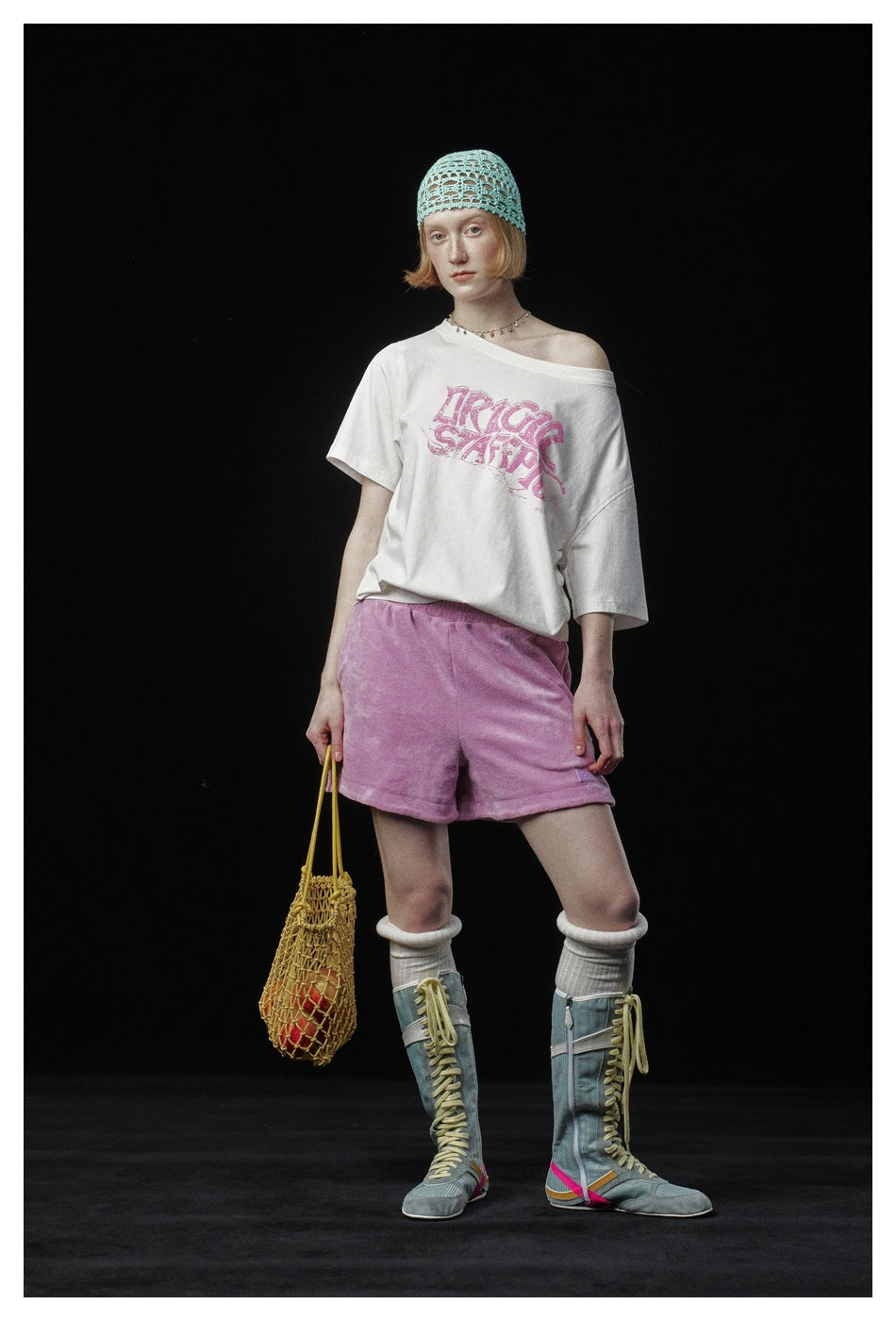 Model wearing an off-white slouchy graphic T-shirt, pink shorts, and a turquoise open-knit cap.