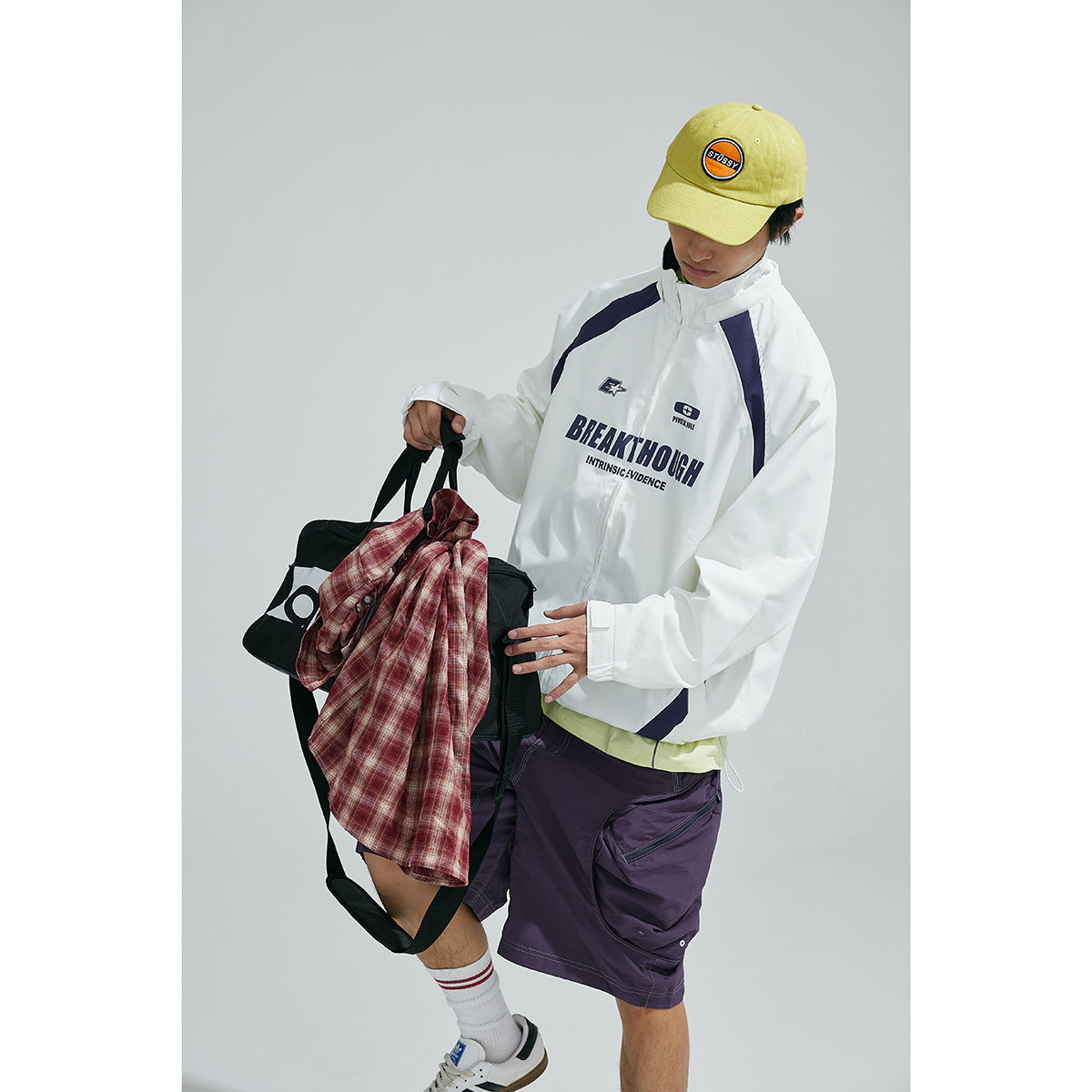Model wearing a white hooded jacket with navy panels and chest graphics, purple shorts, and a yellow cap.