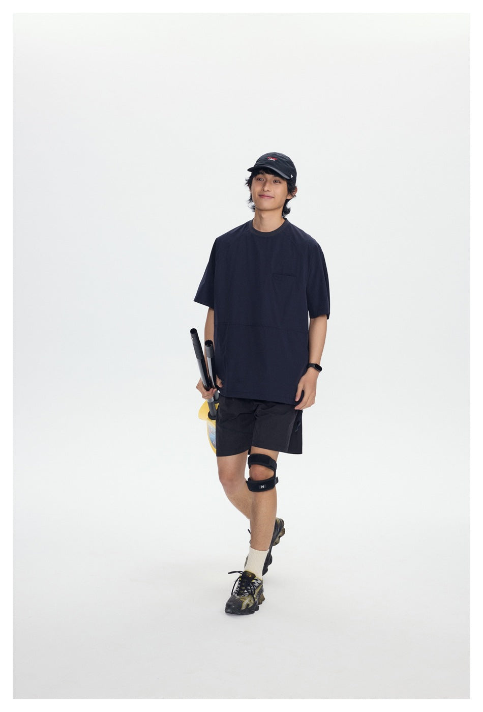 Model in a navy short-sleeve shirt and black shorts, standing with folded paddle equipment against a white background.