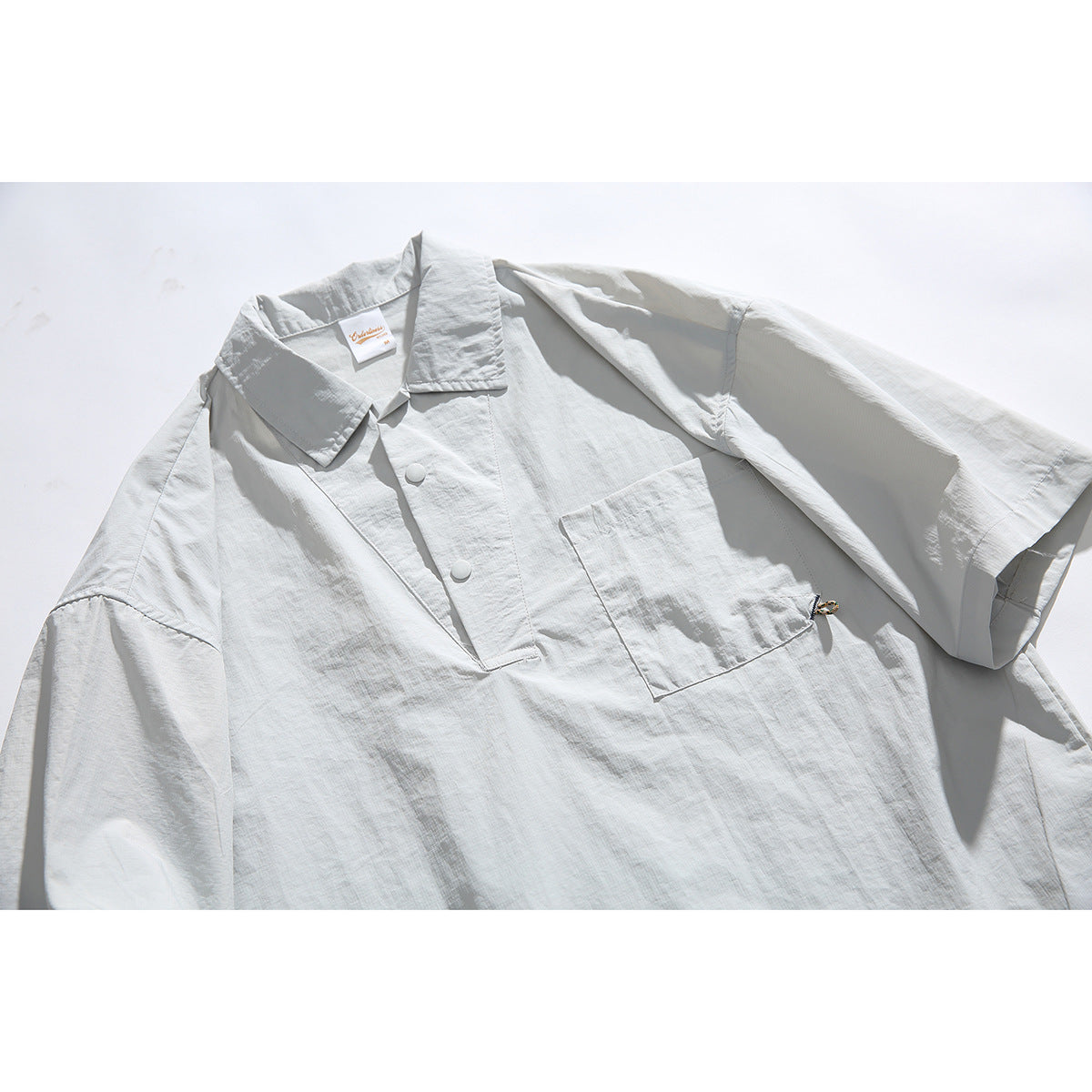 Close-up of a pale gray short-sleeve pullover shirt with a pointed collar, snap placket, chest pocket and side drawcord.