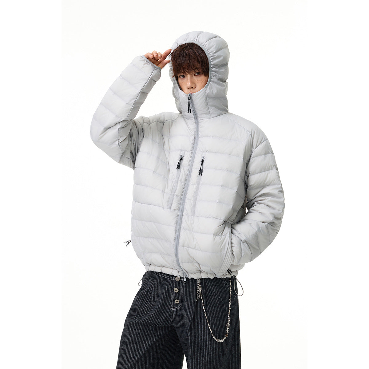 Model wearing a light gray hooded puffer jacket zipped high, with horizontal quilting and two chest zip pockets.