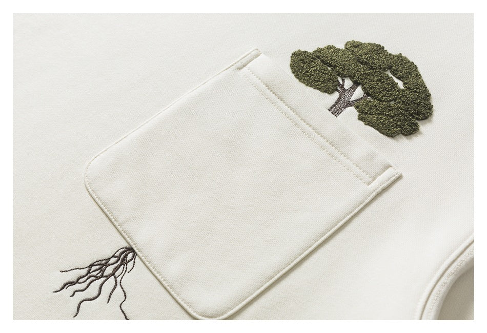 Close-up of a cream sweatshirt’s chest pocket with embroidered tree canopy and roots.