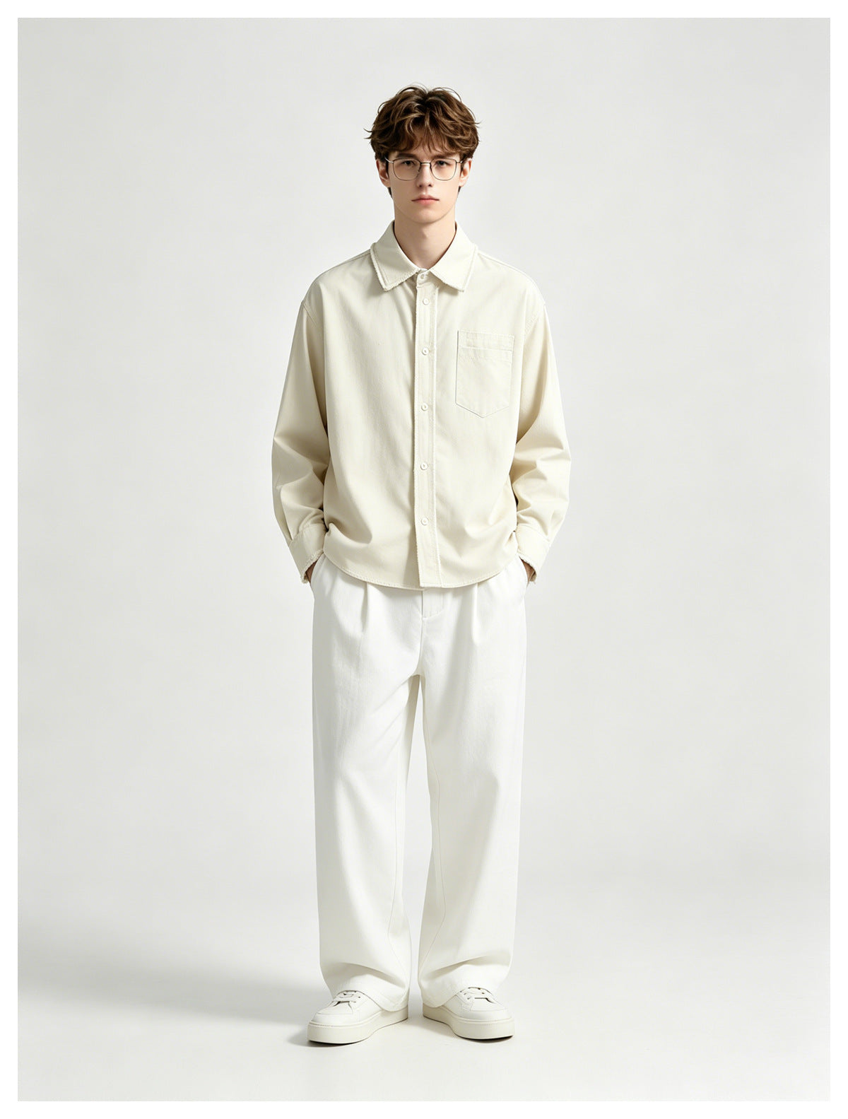 Model standing with hands in pockets wearing a beige frayed-edge button-up shirt, white trousers and white sneakers