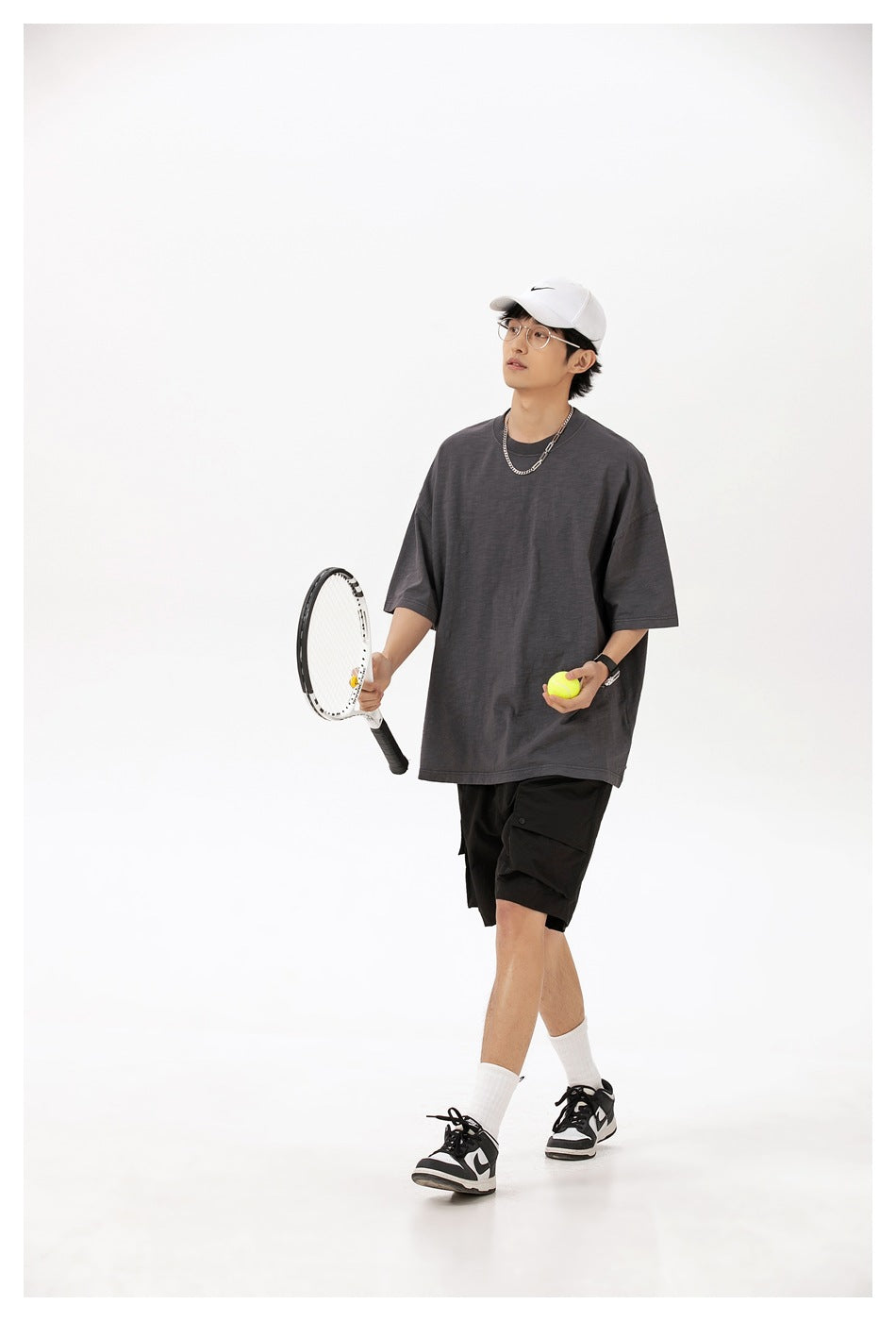 Model wearing an oversized charcoal gray T-shirt and black cargo shorts, holding a tennis racket and ball.