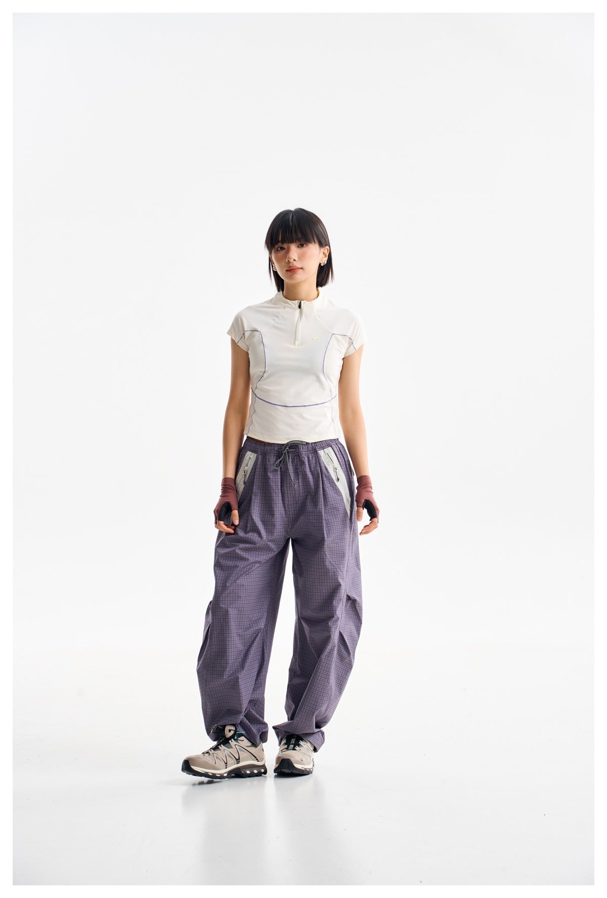 Model wearing a white short-sleeve quarter-zip top with purple seam details and loose purple plaid trousers.