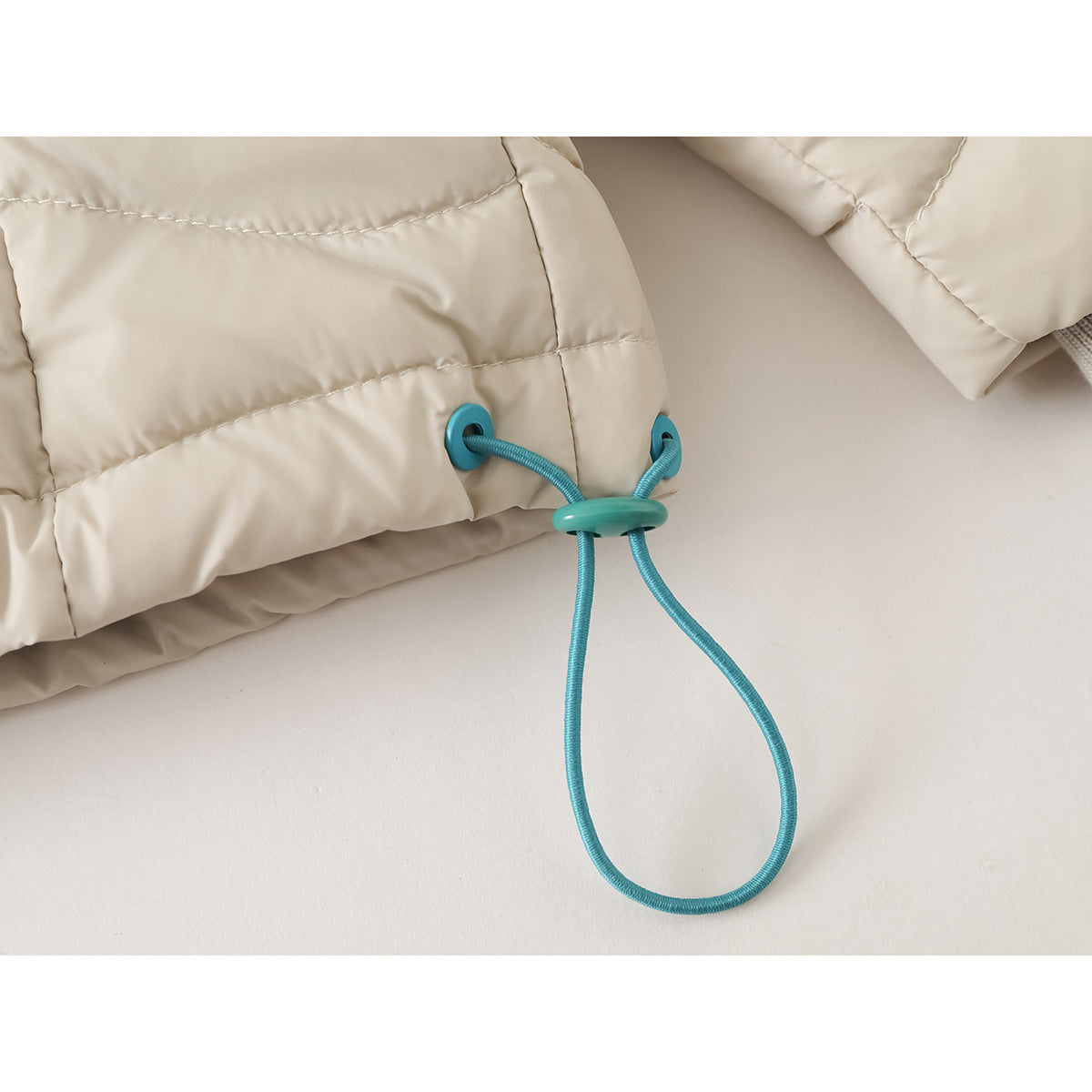 Close-up of a teal adjustable drawcord and toggle at the hem of an ivory quilted jacket.