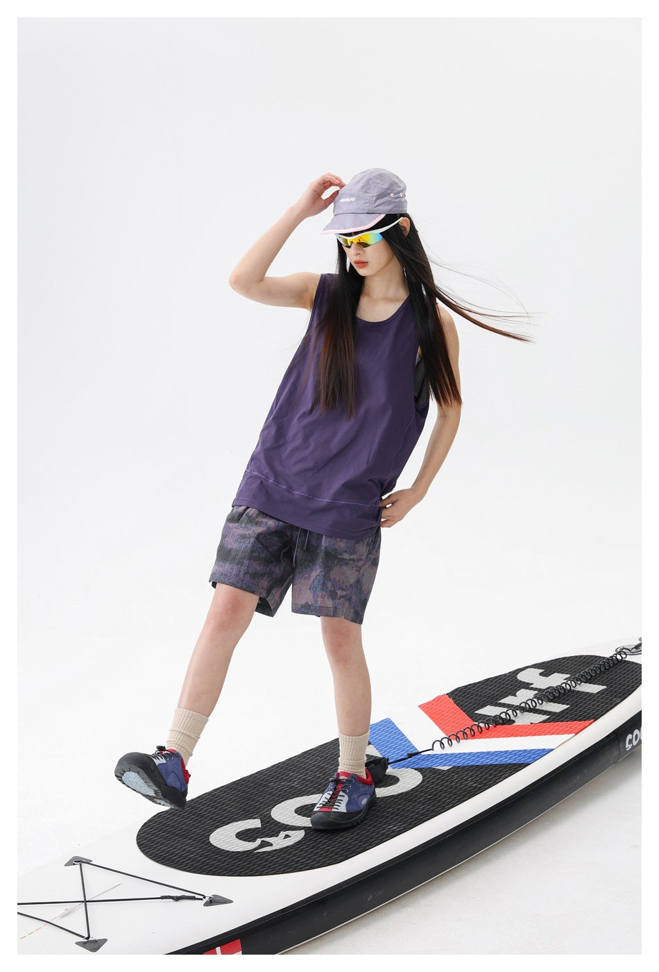 Model in a purple sleeveless tank and patterned dark shorts, standing on a paddleboard.