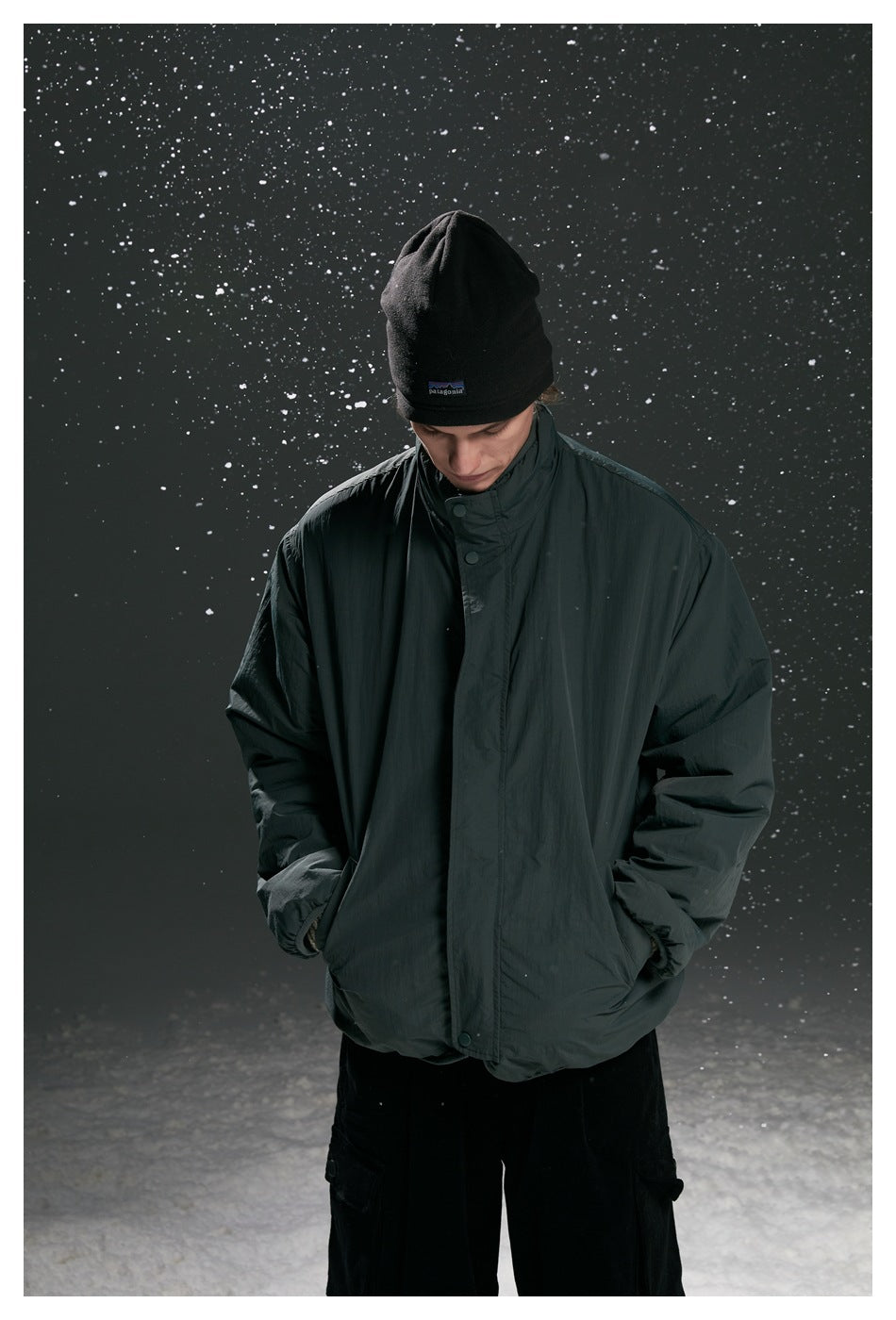 Model looking down in a dark charcoal high-collar jacket with snap-front closure and black beanie.