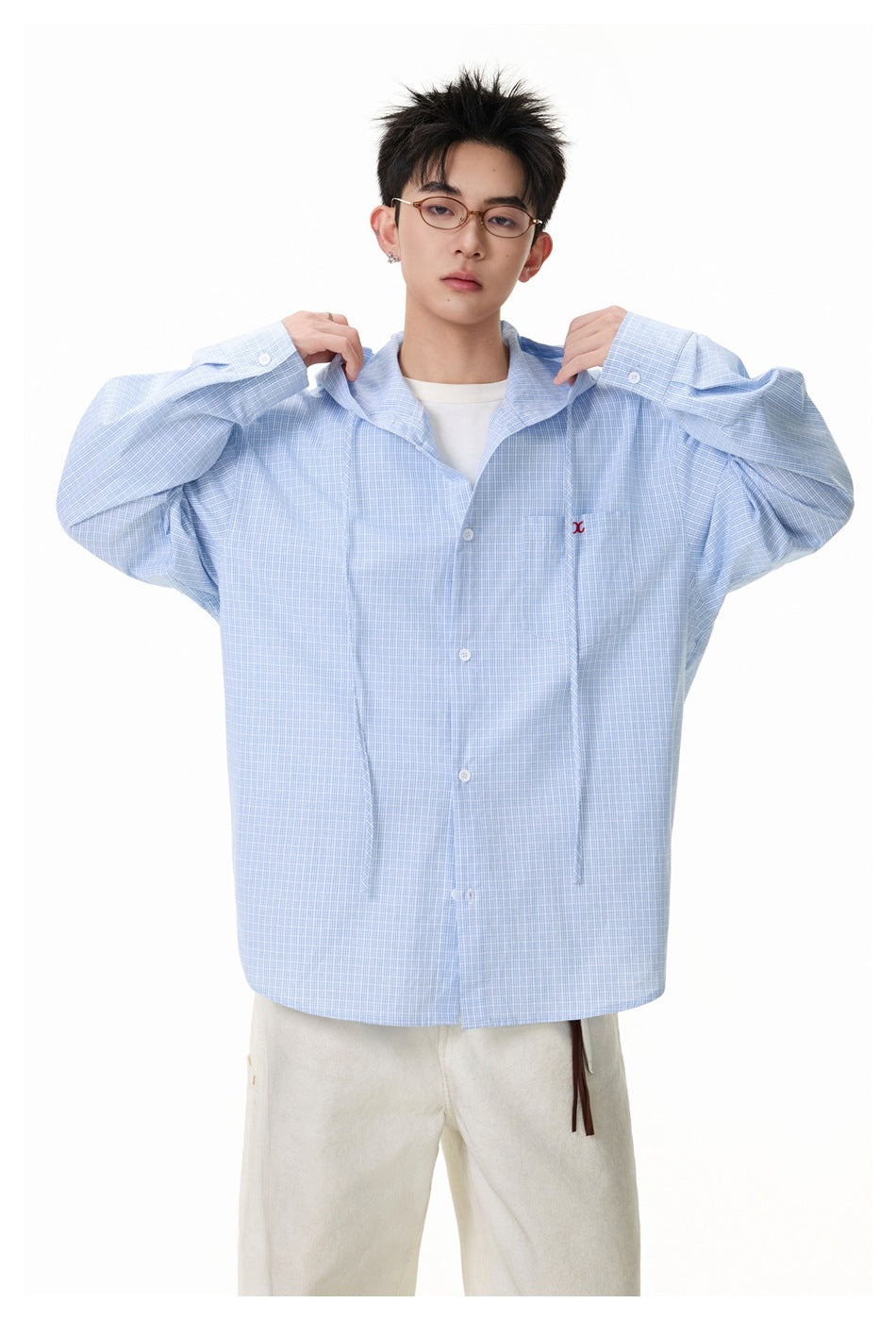 Model holds the hood edges of an oversized light blue checked button-up shirt, shown with cream trousers.