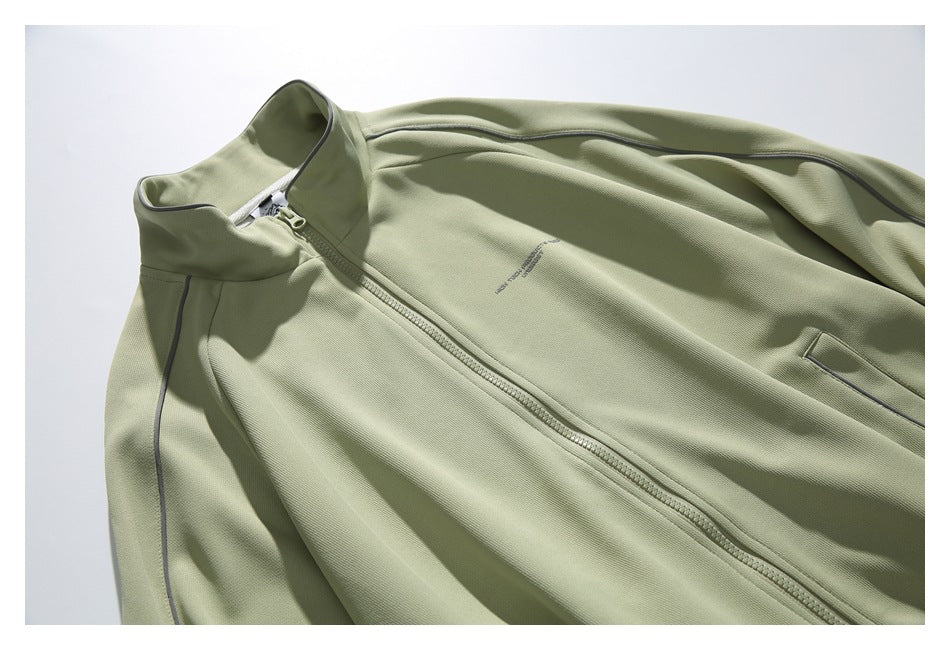 Close-up of the sage-green jacket front showing its zip, stand collar, dark piping, pocket, and small chest lettering.
