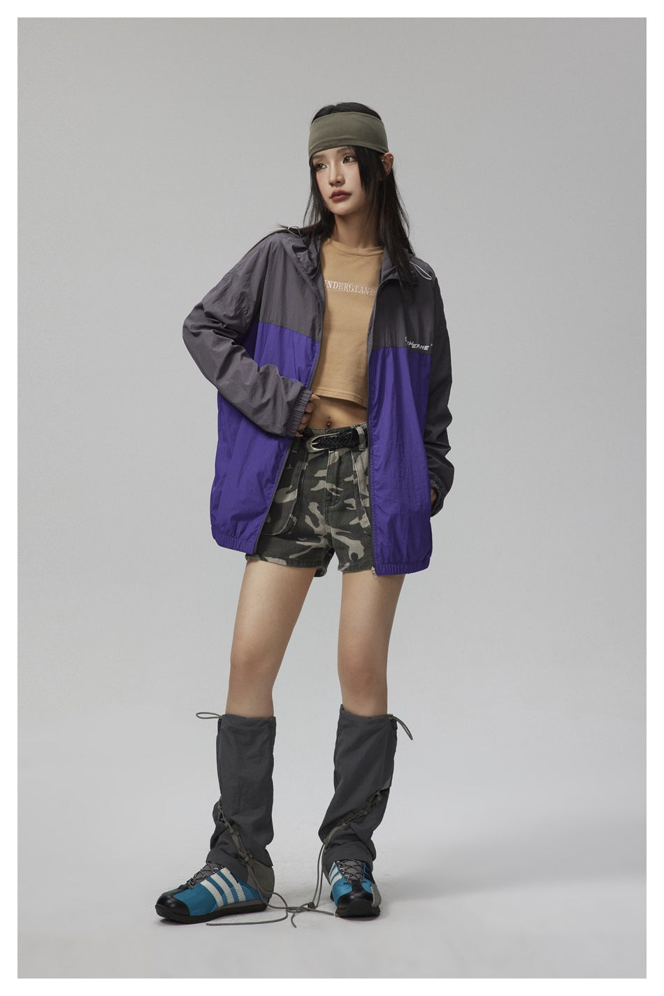Model in the charcoal and purple zip jacket, angled pose with one hand in pocket, over a tan top and camouflage shorts