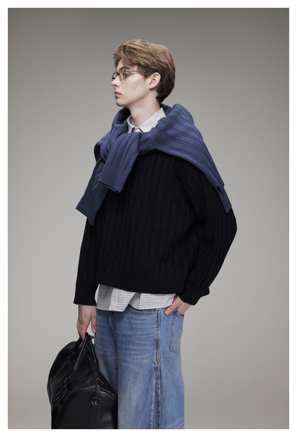 Side view of a model in a black ribbed sweater over a checked shirt, with a blue sweater draped over his shoulders.