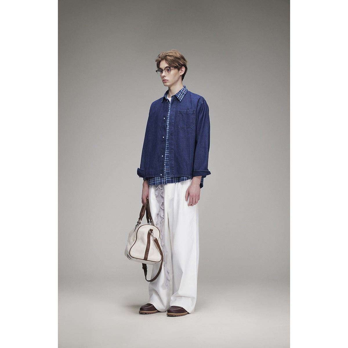 Three-quarter view of a model wearing a navy denim overshirt over a blue plaid shirt and loose white trousers, with a cream