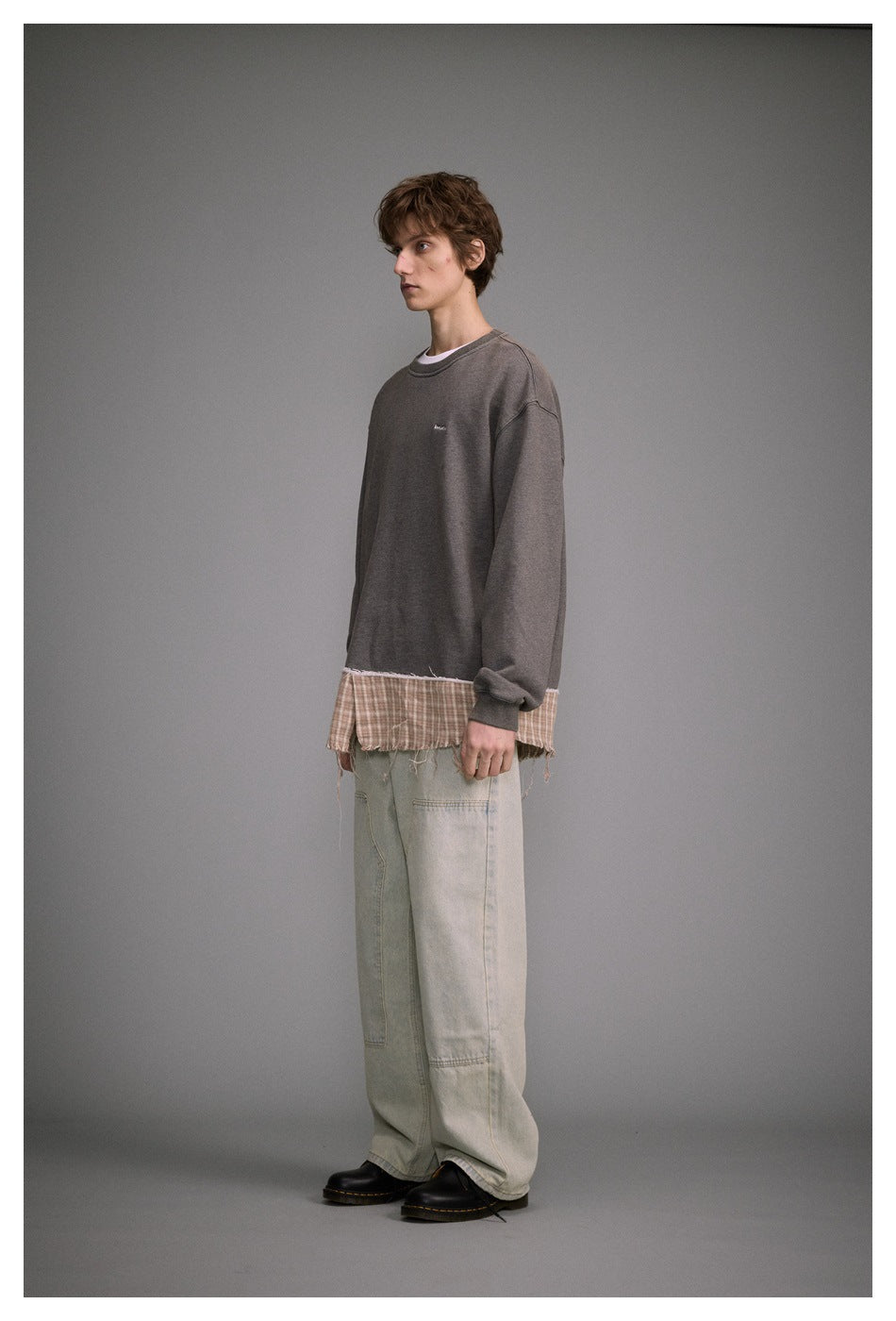 Model in side view wearing gray sweatshirt with plaid frayed hem panel, light wash wide-leg jeans and black shoes