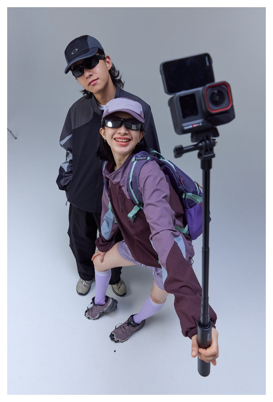 Two people in color-block jackets pose for a selfie; a large action camera partly obscures the foreground.