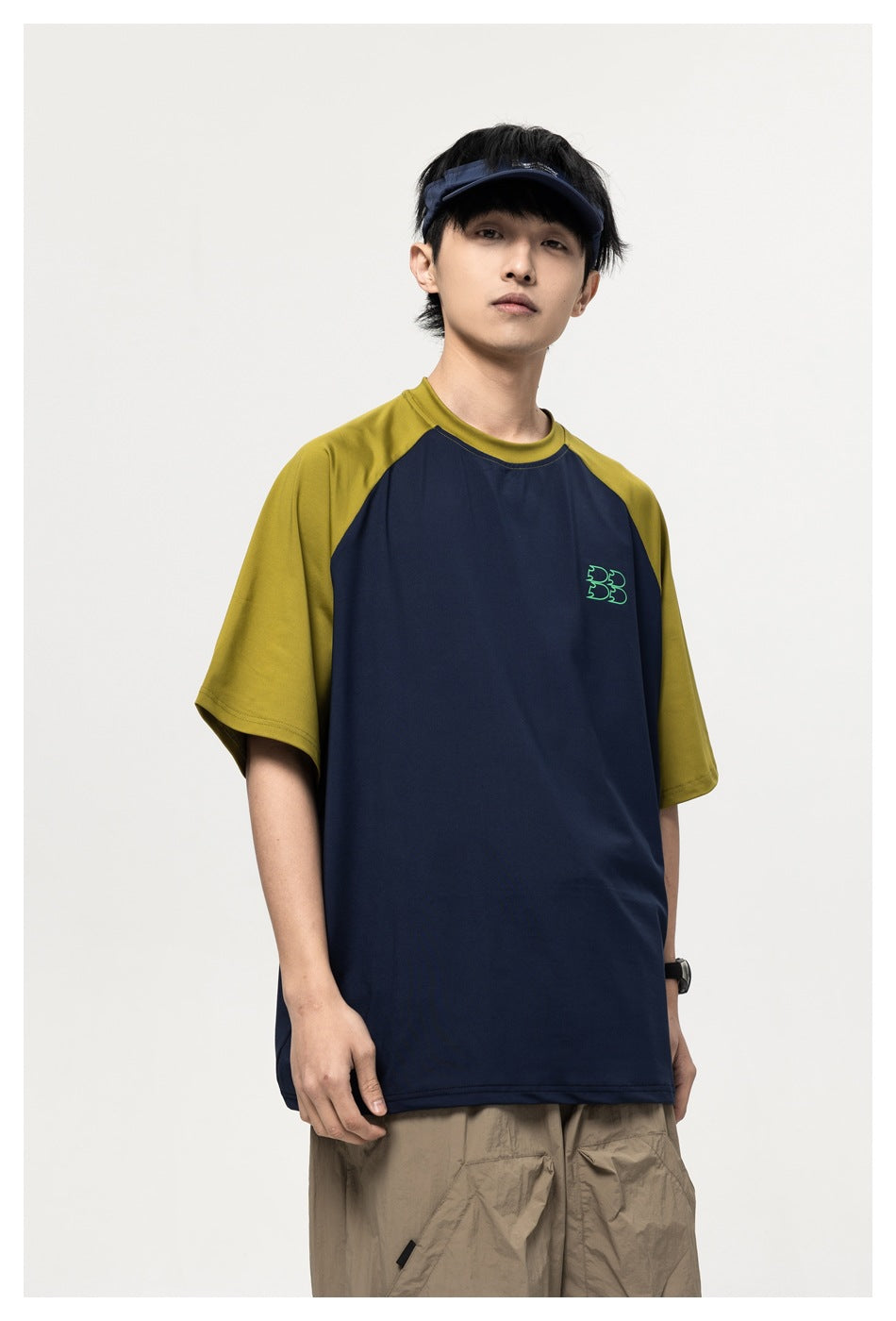 Model in navy raglan T-shirt with olive sleeves and small green outline logo on chest, with visor and khaki pants
