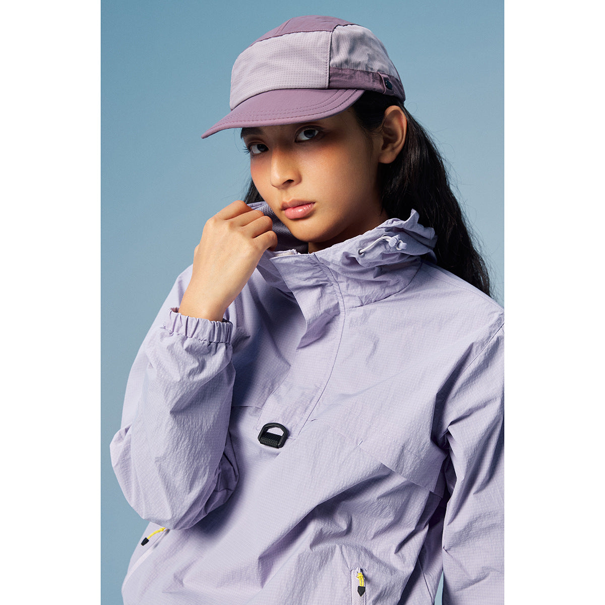 Model wearing a pale lavender hooded pullover jacket and matching-panel cap against a blue backdrop.