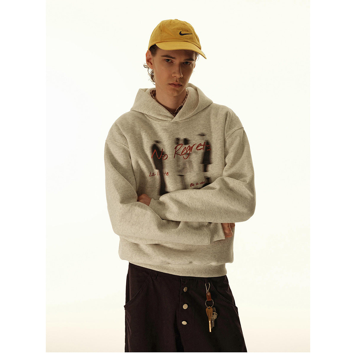 Model wearing a light gray hooded sweatshirt with red “No Regrets” artwork and dark trousers.