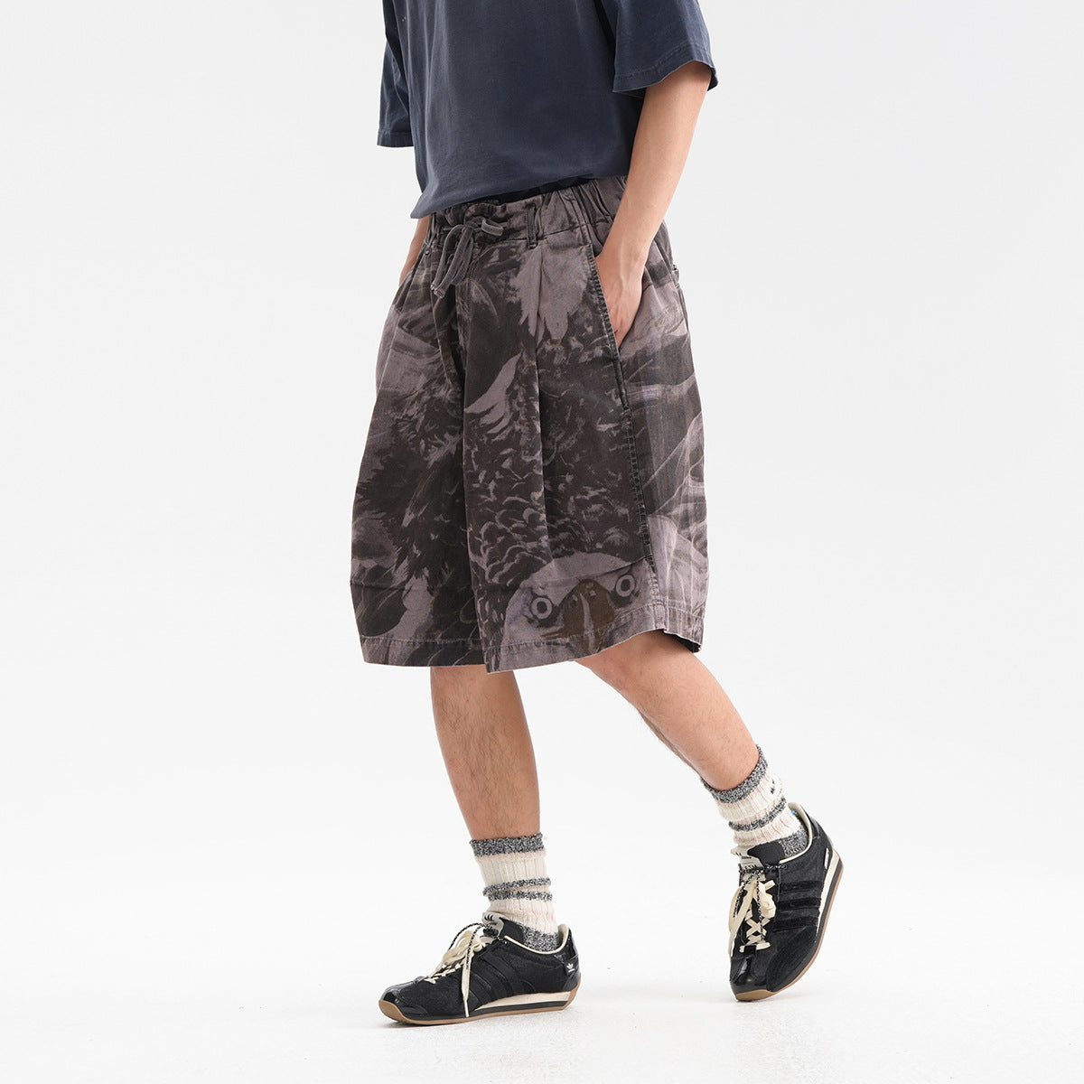Model wearing loose charcoal-gray graphic shorts with an elastic drawstring waist and wide legs, shown from the front at an