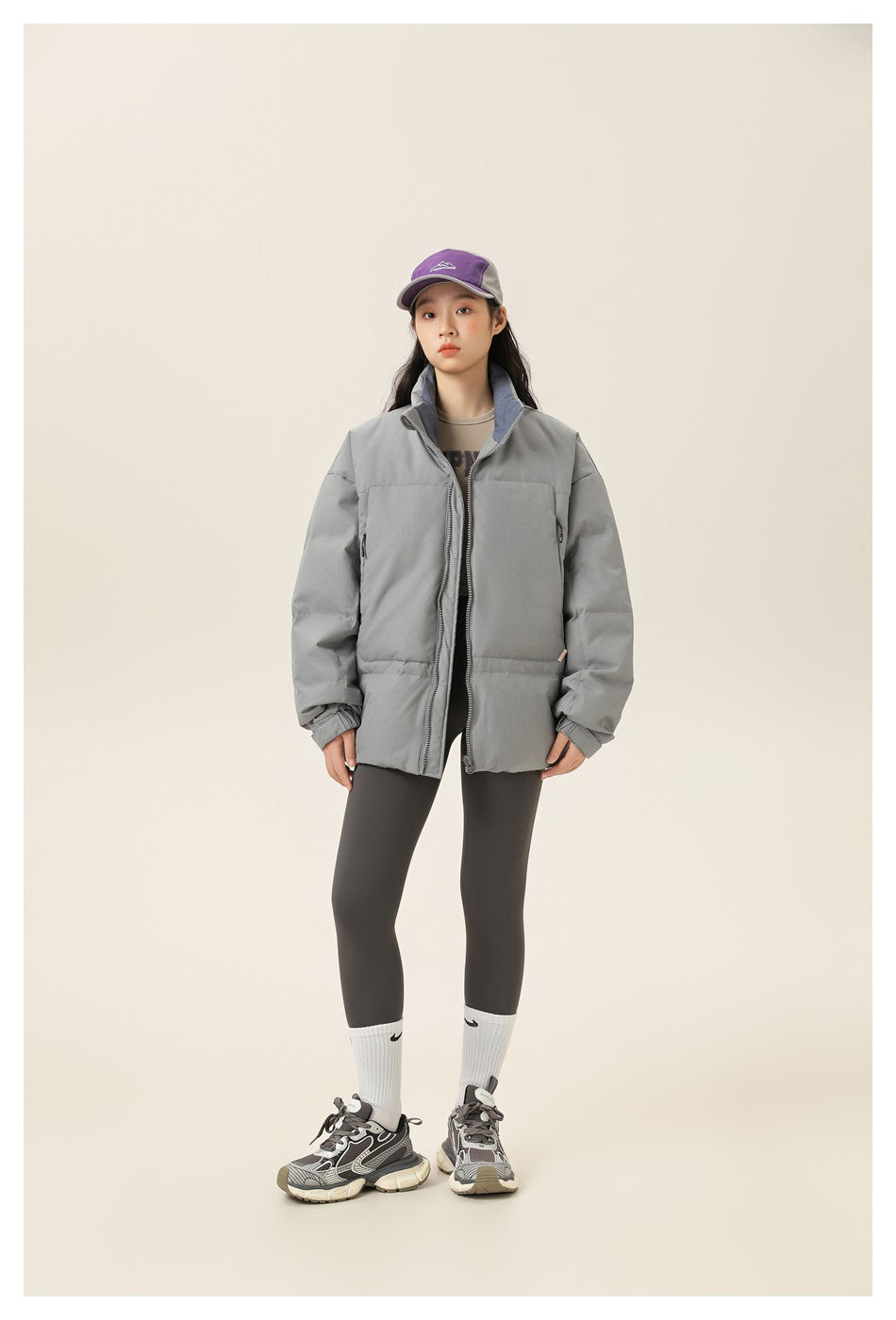 Model wearing gray puffer jacket with stand collar, open front zipper, dark leggings, purple cap and sneakers