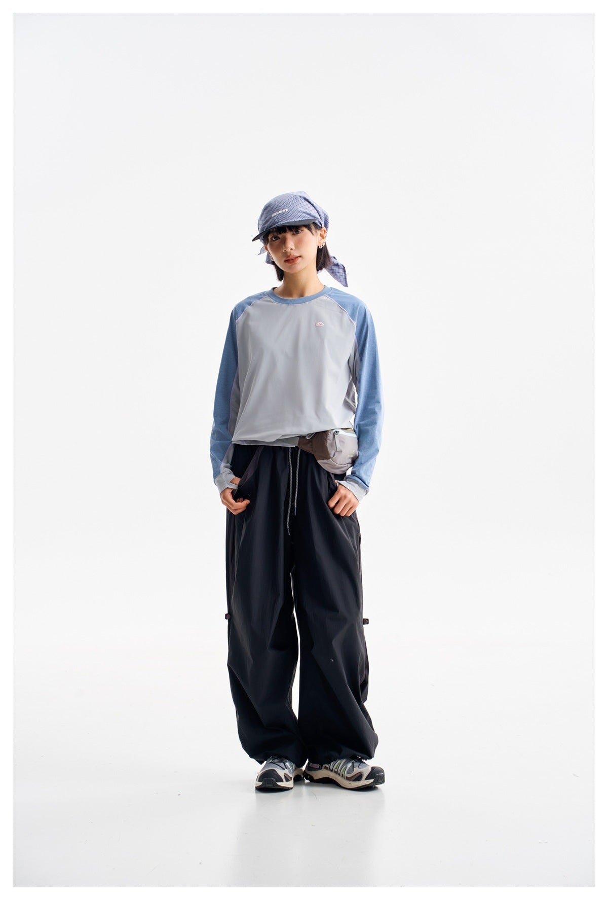 Model facing forward in a light gray top with blue sleeves and loose dark pants, wearing a plaid cap.
