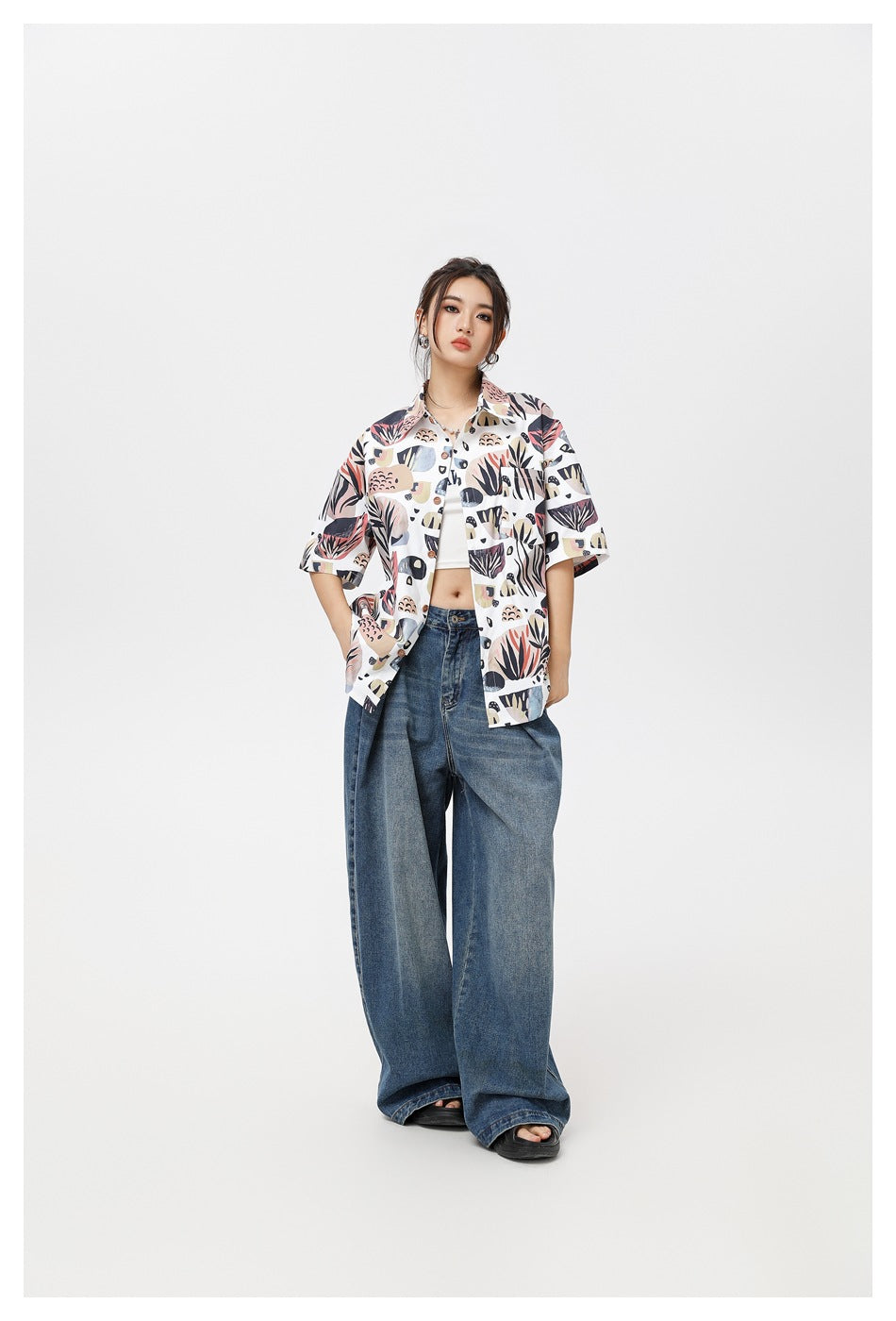 Full-length model wearing open botanical-print short-sleeve shirt with white top, wide-leg jeans and sandals