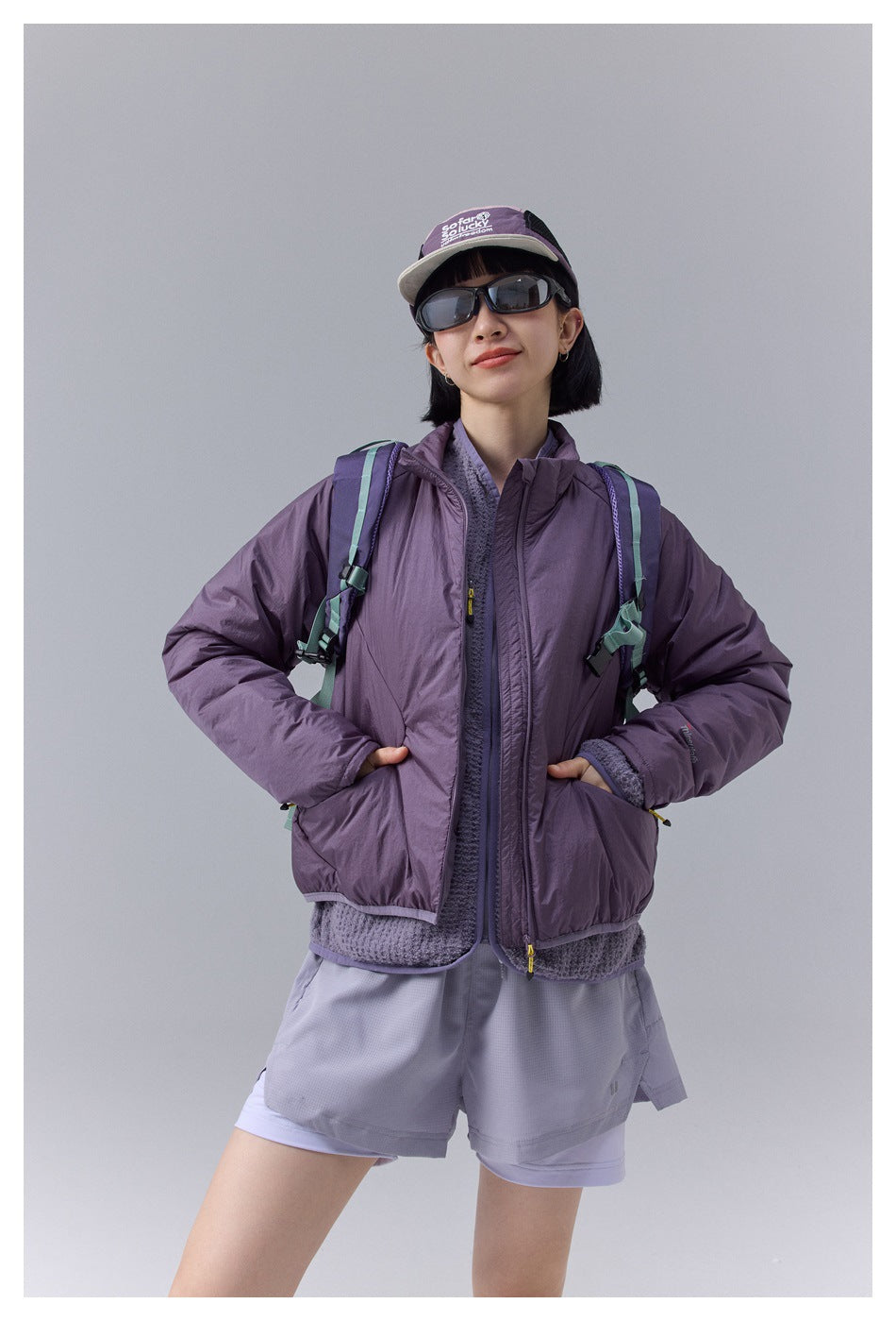 Woman wearing a purple padded stand-collar jacket over a knit layer with shorts, cap, sunglasses and backpack