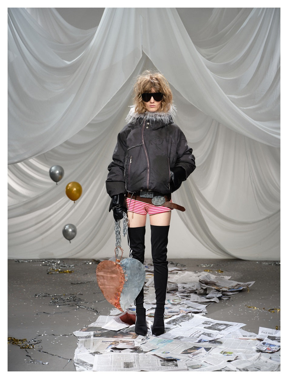 Model wearing the dark hooded jacket with gray trim, striped shorts, belt and tall black boots against white drapery
