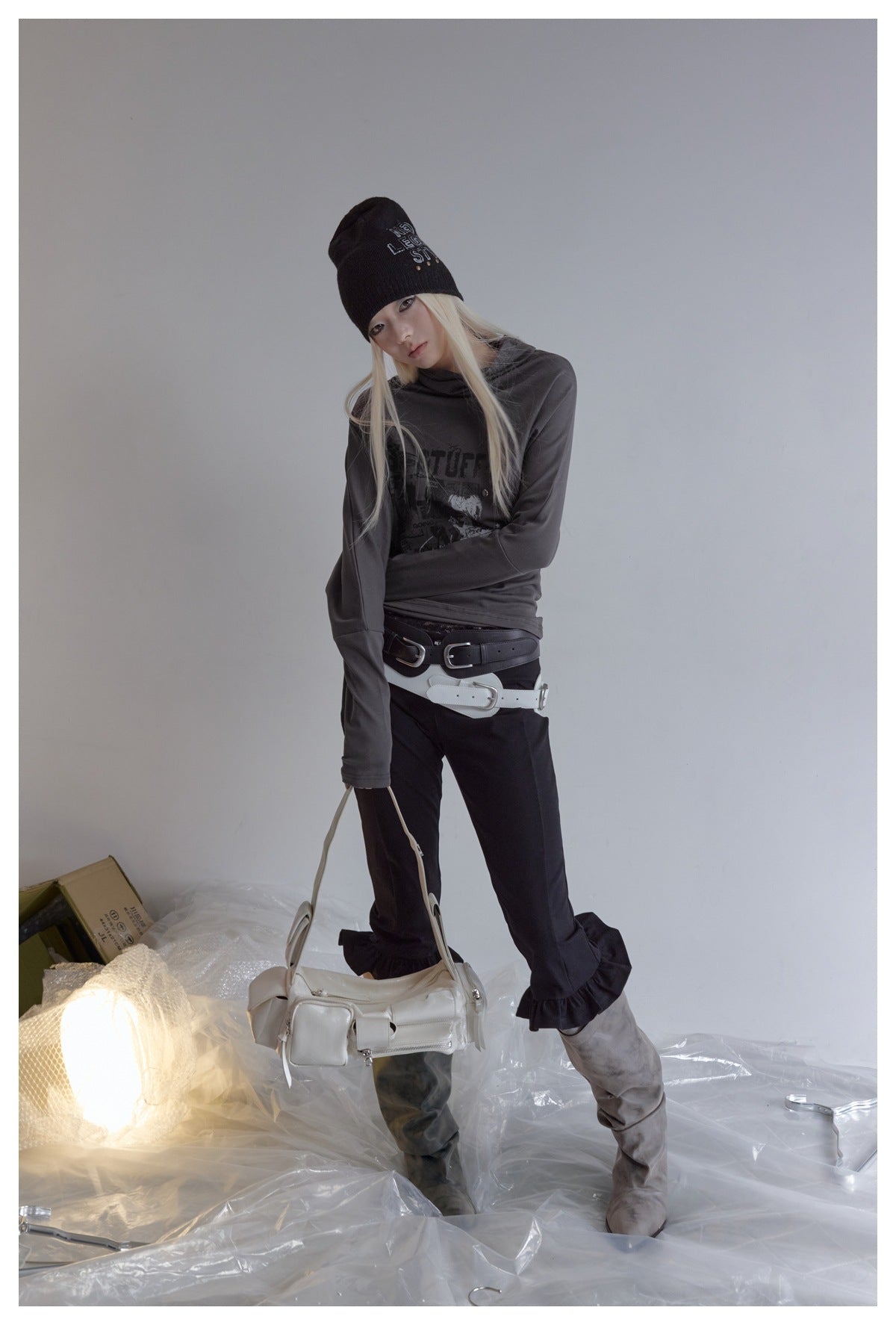 Model wearing dark gray graphic high-neck top with black beanie, dark flared pants and gray boots, holding a white bag