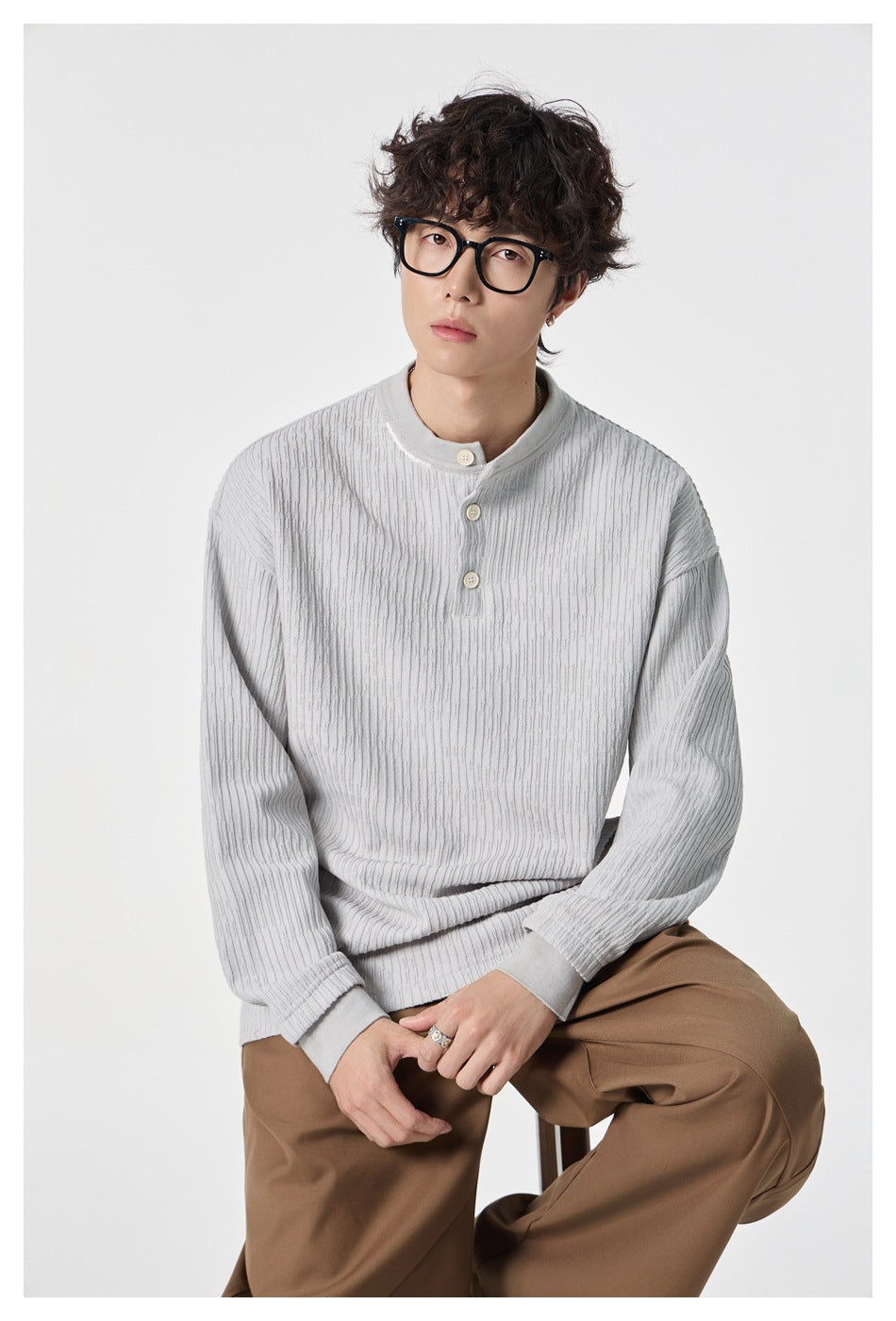 Seated model wearing a pale gray textured long-sleeve top with a three-button collar and brown trousers.