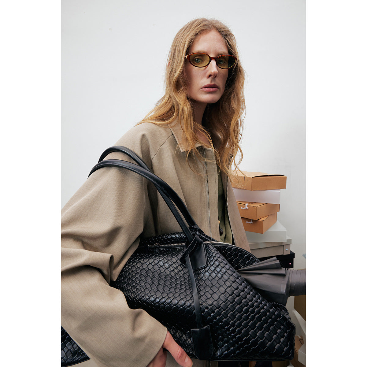 Close view of a model wearing a taupe zip-front jacket, with a large black woven handbag in the foreground.