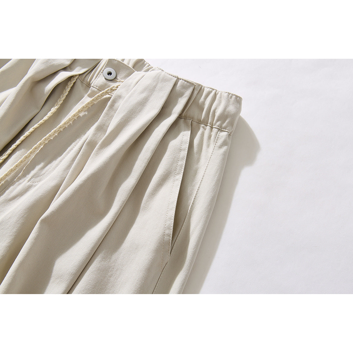Angled close-up of ivory pants showing the gathered waistband, button, pleats and side pocket seam.