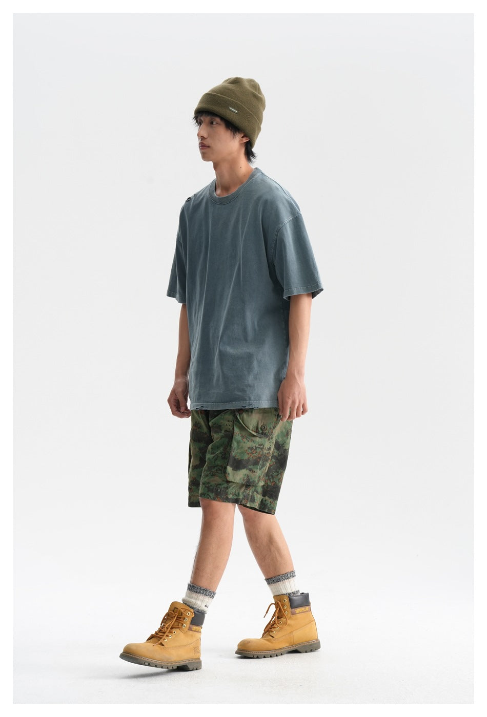 Model wearing a faded blue-gray T-shirt, green camouflage cargo shorts, olive beanie, and tan boots.