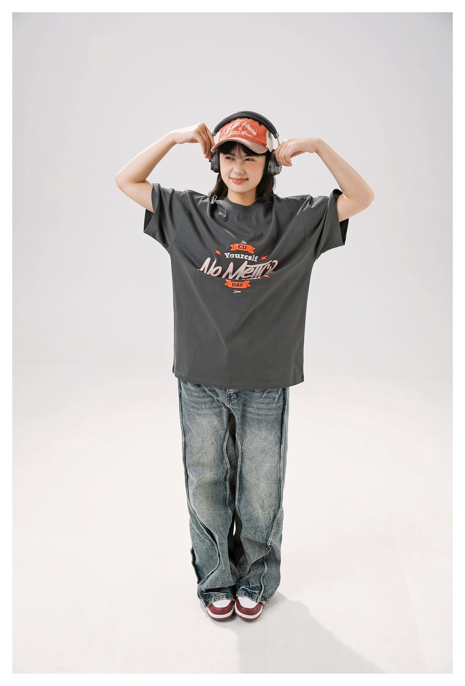 Model wearing a charcoal gray oversized graphic T-shirt with loose faded jeans, holding headphones.