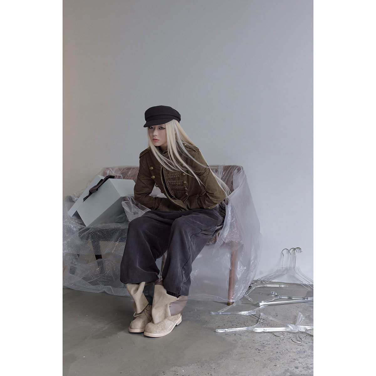 Model seated in an olive-brown military-style jacket with brass buttons, dark wide-leg trousers, and a black cap.