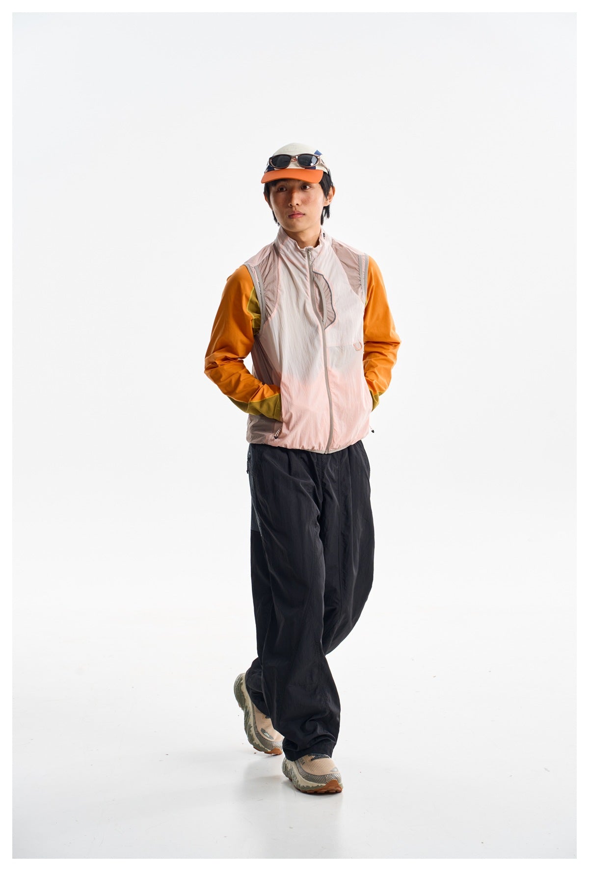 Model wearing loose black pants and a pale pink zip vest over an orange top, walking toward the camera.