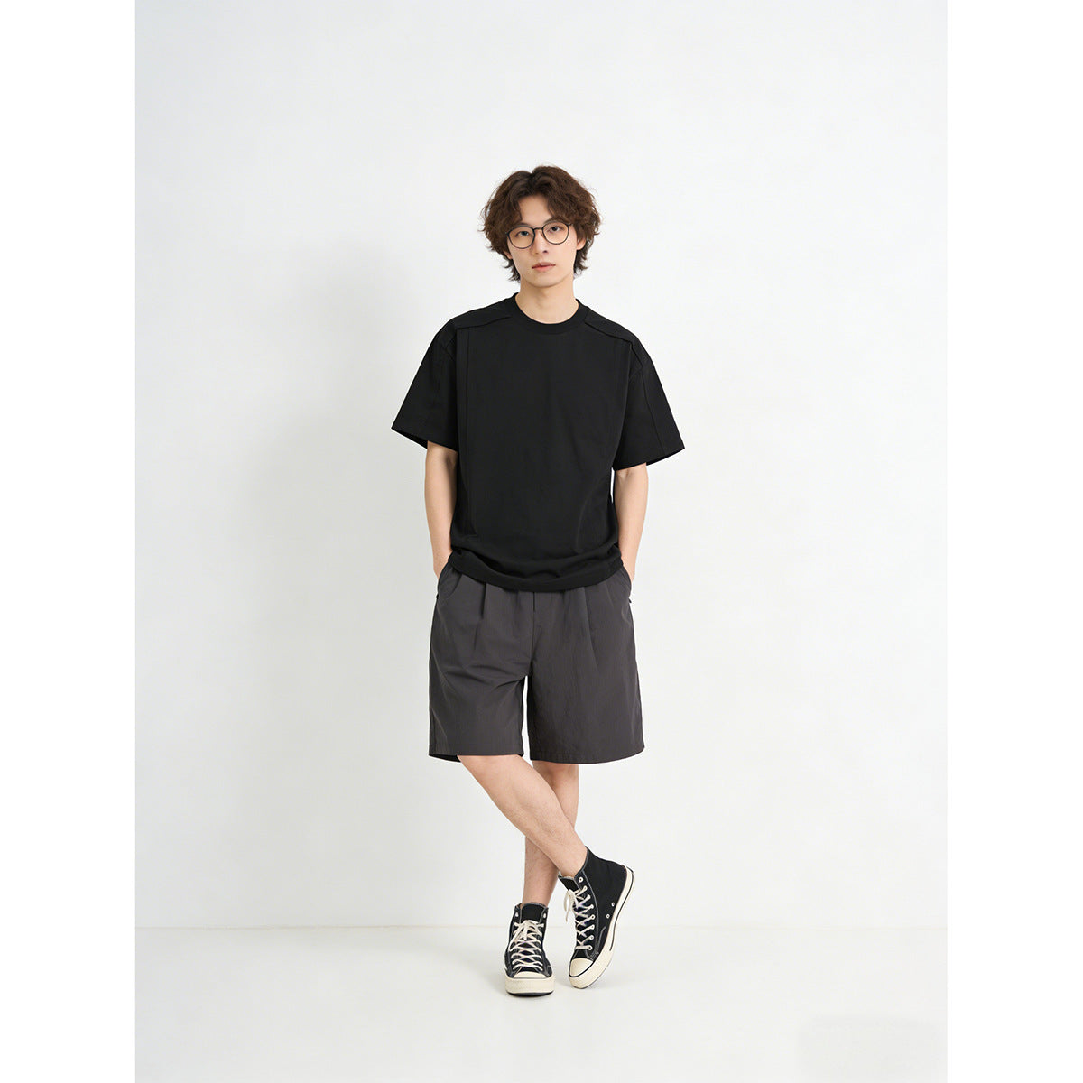 Model wearing a plain black short-sleeve T-shirt with charcoal shorts and black high-top sneakers, full-length front view.