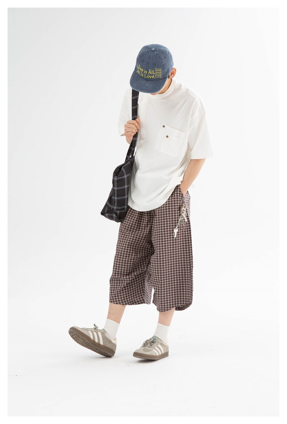 Model wearing a white short-sleeve shirt with a buttoned chest pocket and brown plaid shorts, carrying a dark plaid bag.