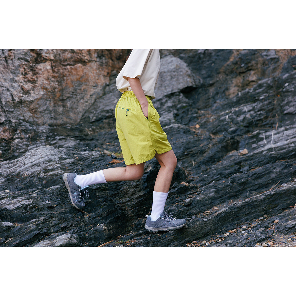 Cropped model stepping across rocks in bright lime elastic-waist shorts and a white top.