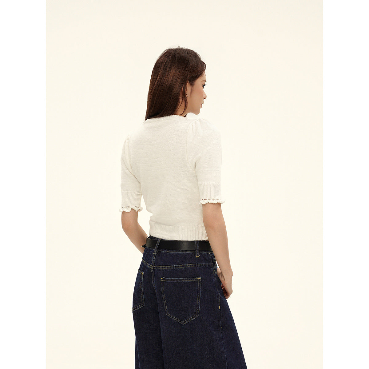 Rear view of a model wearing an ivory knit top with scalloped sleeve trim and dark wide-leg jeans.