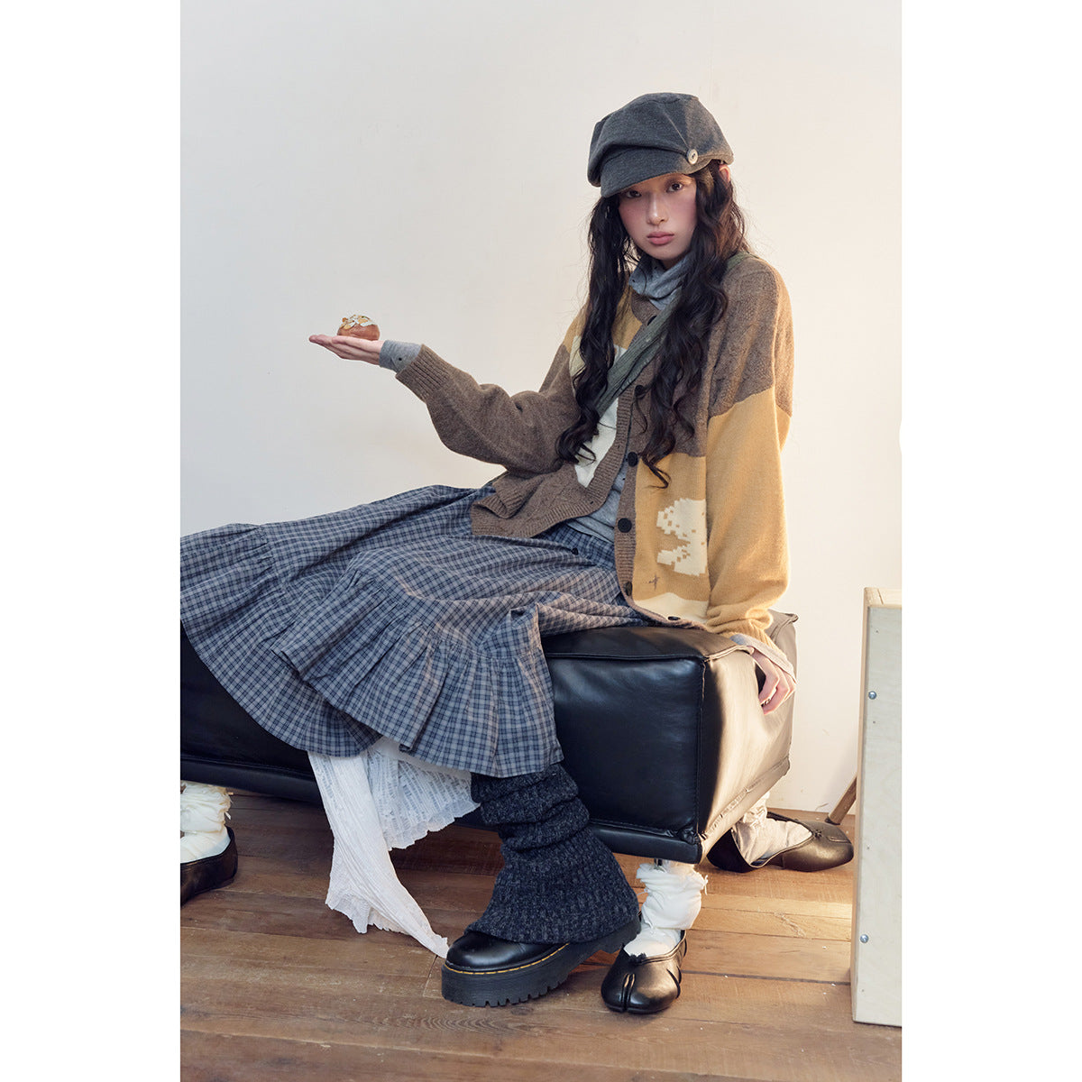 Seated model wearing a brown and mustard cardigan with a blue plaid skirt, gray cap, and dark leg warmers.