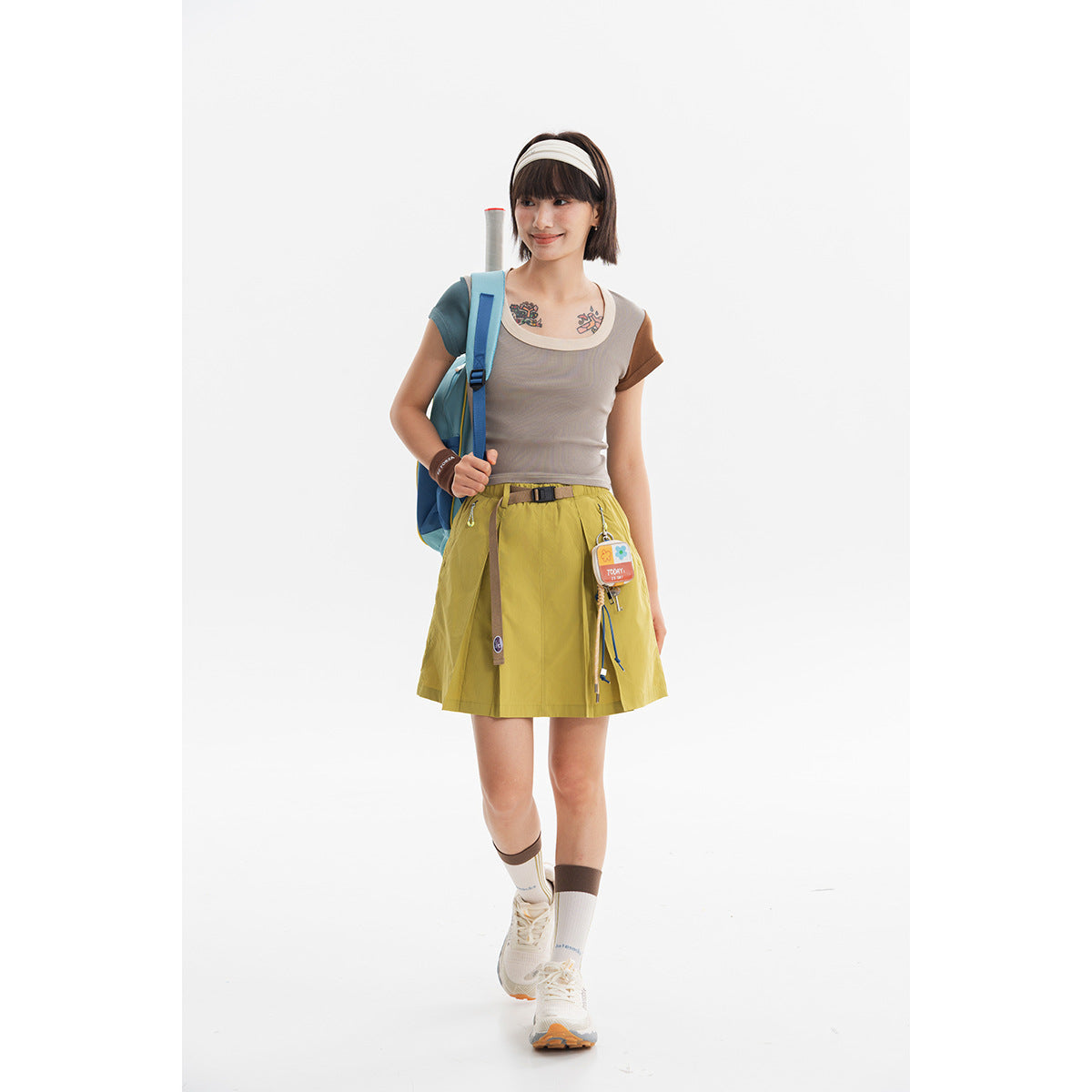 Model wearing a taupe short-sleeve top with contrasting sleeves and a mustard yellow tennis skirt.