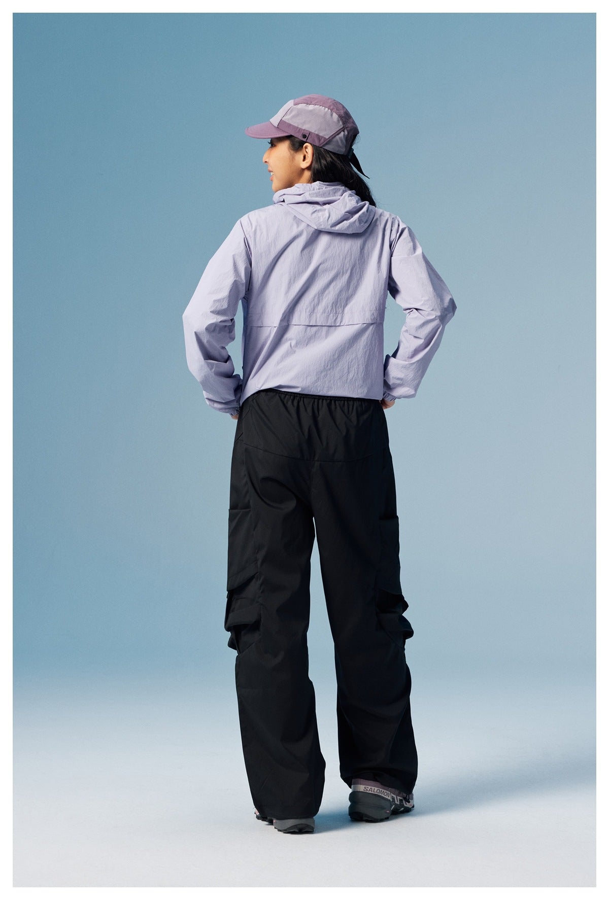 Rear view of a model in a light lavender hooded jacket and loose black pants, wearing a matching cap.