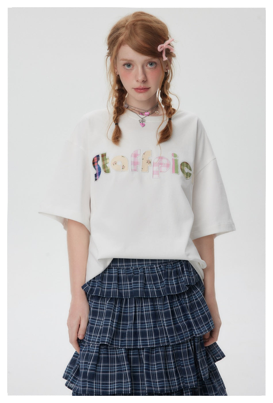 Front view of a model in an oversized white T-shirt with colorful patchwork lettering and a navy plaid ruffle skirt.