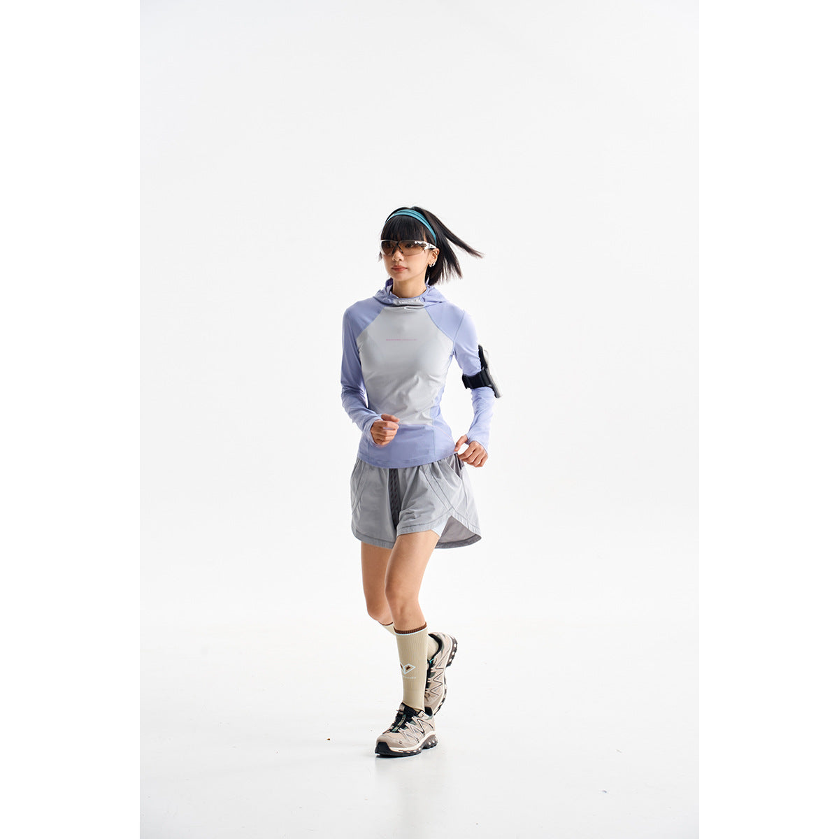 Model in a pale gray and lavender hooded top with gray running shorts and sneakers.