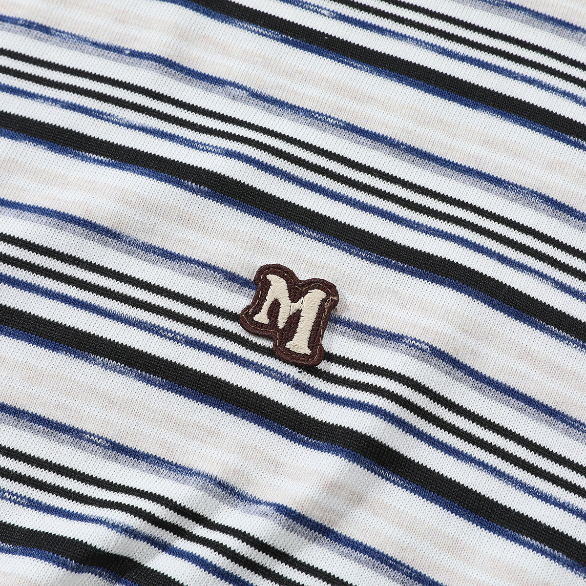 Close-up of a small embroidered M patch on white knit fabric with black, navy, and gray stripes.