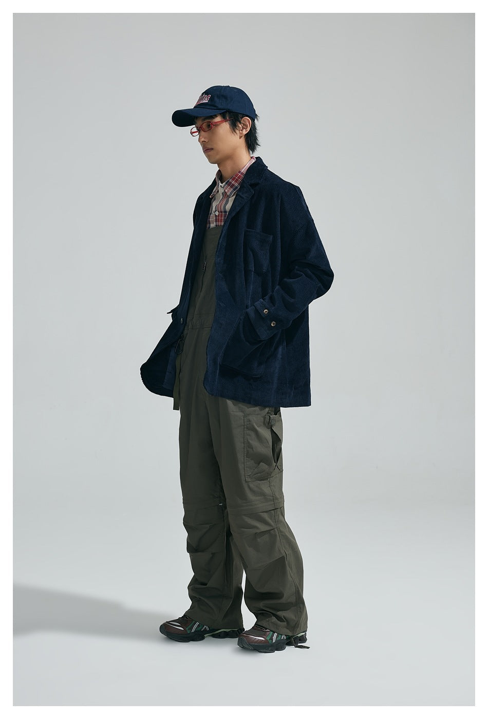 Model wearing navy corduroy jacket over plaid shirt with olive overalls and a navy cap, side view